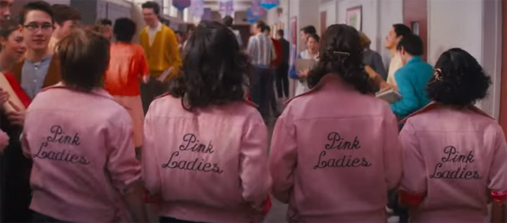 Four high school girls walking the high school halls, backs to camera with Pink Ladies emblazoned on their jackets