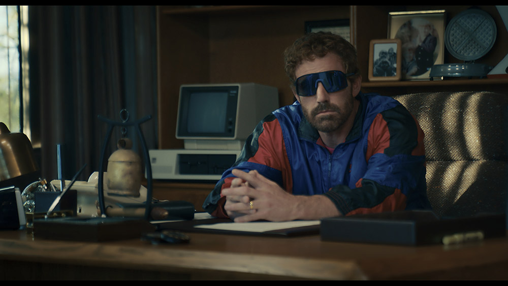 Man in sunglasses and 80's style curly perm and tracksuit sitting at on office desk