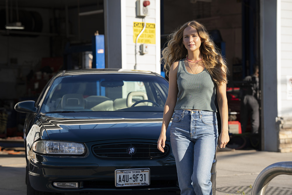 A woman in jeans and grey tank top walking in front of a car